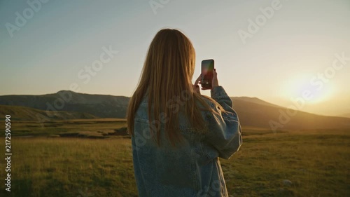 A person takes a picture with a smartphone while standing in a green field during sunset