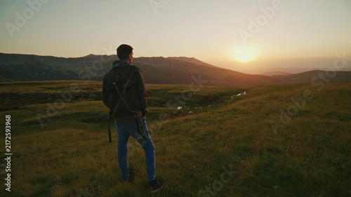 Man stands on grassy hill watching sunset over mountains in the background with a stream flowing through the valley during late evening