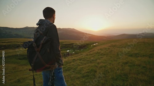 Young man watches the sunset while standing on a grassy hillside near a stream in an open valley