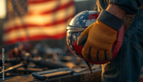 A construction worker holding a helmet with an American flag pattern at a job site.