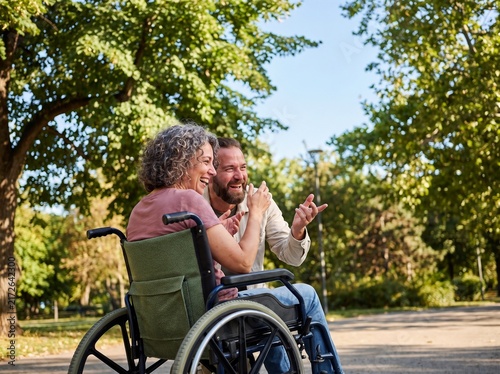Cheerful mature woman in a wheelchair laughing with a male friend on a sunny park alley, joyful disability lifestyle, supportive relationship and accessible outdoor mobility