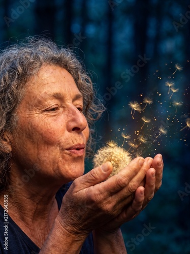 Profile portrait of a senior woman blowing a dandelion with magically glowing golden seeds flying in a dark forest, hope, imagination and active aging lifestyle concept