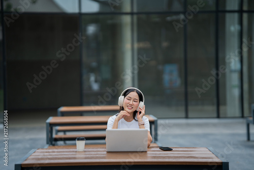 Картината върху платно A smiling young woman wearing headphones works remotely on her laptop, studying