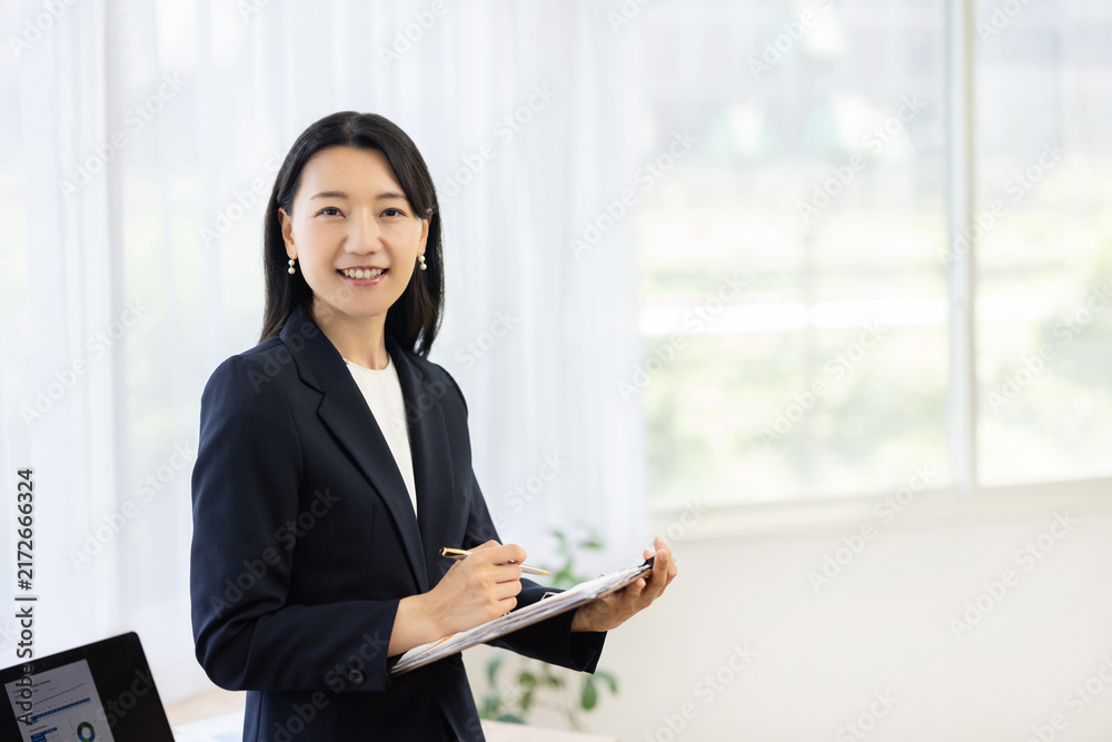 Obraz premium Young Japanese businesswoman holding a clipboard and smiling at the camera by a bright window