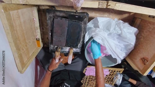 Person wears gloves, works with pink wax. Hands press wax into wooden mold. Wax melts in heated, rustic container. Gloved hands guide the process carefully. Traditional craft captured in close-up view