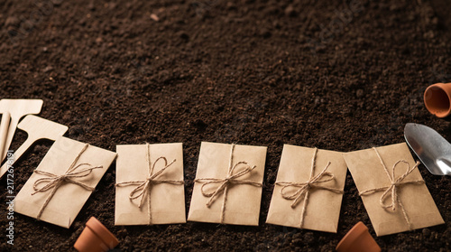 Top view of rustic craft paper seed packets tied with twine arranged on dark fertile soil with gardening tools for spring planting preparation and organic vegetable garden concept