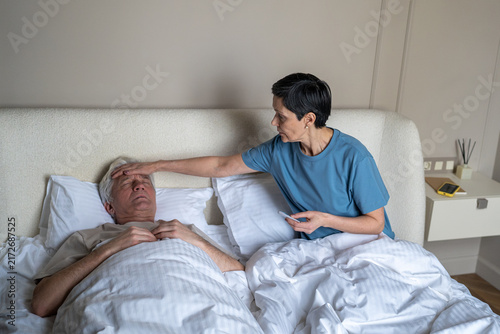 Photography Anxious mature wife taking temperature of ill feverish husband in bed with thermometer