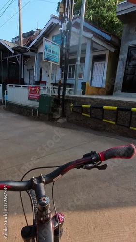 Vertical POV video of Bicyclist rides through quiet village streets. Sunlight casts warm glow on narrow roads. Simple houses line the path with greenery. Handlebars frame view as cyclist moves forward