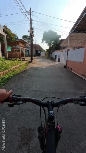 Vertical POV video of Bicyclist rides through quiet village streets. Sunlight casts warm glow on narrow roads. Simple houses line the path with greenery. Handlebars frame view as cyclist moves forward