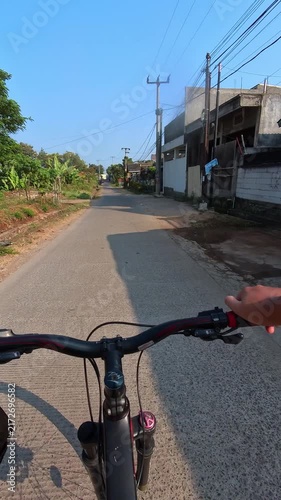 POV Video of Bicycle handlebars frame a quiet village street. Clear blue sky above a calm, sunlit road. Hand grips handlebars, suggest motion and purpose. Cables stretch overhead, connecting homes and