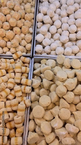 Vertical Video of Various frozen meat and seafood meatballs in metal trays at a market stall. Baked goods display textures, shapes, and colors. Golden, white, and brown items fill each section. 