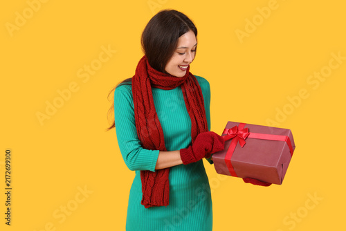 Beautiful young Asian woman in warm scarf with gift box on yellow background