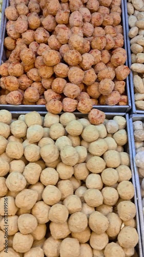 Vertical Video of Various frozen meat and seafood meatballs in metal trays at a market stall. Baked goods display textures, shapes, and colors. Golden, white, and brown items fill each section. 