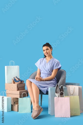 Young woman with shoe boxes and shopping bags sitting in armchair on blue bac...