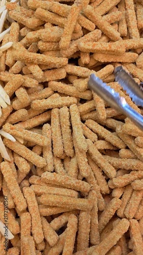 Video of Golden, crispy chicken nugget sticks fill the frame. Tongs grasp one stick, ready for serving. Texture looks crunchy, perfectly fried. Hand holds tongs, suggests a snack moment. Image evokes 