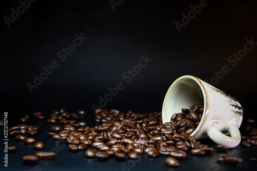 Принти на полотні Roasted coffee beans spilling from a tipped ceramic cup onto a dark surface, photographed against a black background with generous copy space