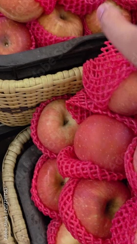 Vertical video of Hand selects a red apple from a pink netted bag. Apples glow with fresh, vibrant red skin. Basket holds more apples in soft mesh. Fruit displays warm, inviting market appeal. 4K POV 