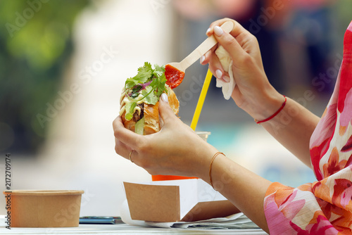 Elegant woman with manicured hands and colorful floral outfit drizzling red sauce onto a loaded street food sandwich, savoring an unhurried moment at Karlin food festival in Prague, Czech Republic. 