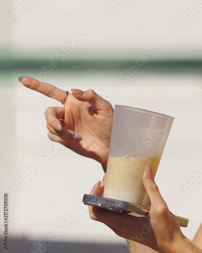 A woman gestures with one hand while holding a chilled iced coffee and her phone in the other, a candid on-the-go moment at the sunny Naplavka farmers market in Prague.
