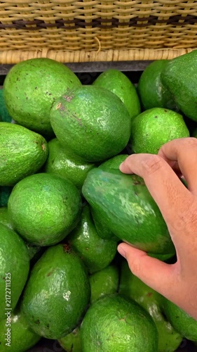 POV Video of Hand selects ripe avocado from market basket. Firm, smooth green fruit gleams under soft light. Market scene suggests fresh produce, natural choice. Hand shows gentle care