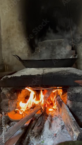 Vertical shot of a traditional wood-fired stove with a steaming pan. Authentic rural kitchen cooking process using burning firewood. Rustic Asian culinary background.