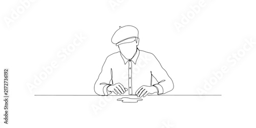 Continuous line drawing of a man wearing a beret cap sitting at a table working with his hands