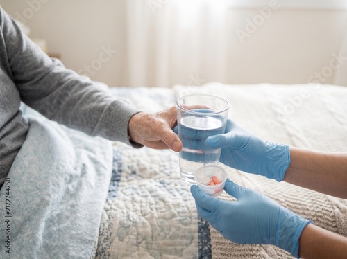 Caregiver in blue gloves offers water and medication to elderly person in bed.