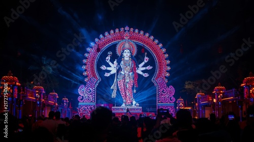 Majestic multiarmed deity idol illuminated by vibrant stage lighting in elaborate nighttime festival setting with illuminated backdrop with captures towering intricately