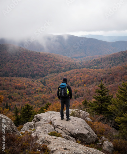 Backpacker male hiker standing on mountain rock peak enjoying view of misty colorful forest valley in autumn