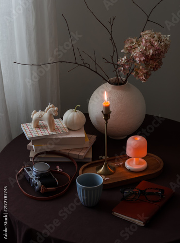 Cozy autumn home interior - a vase with flowers, a stack of books, a camera, candles, and a glass of coffee on a round table with a brown tablecloth in the living room