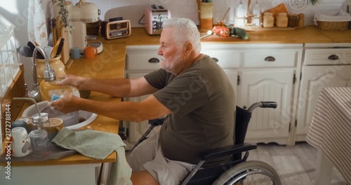 Side view of an independent senior man in a wheelchair washing dishes at the kitchen sink, showing active daily life and self sufficient living at home.