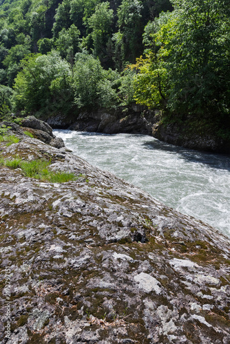 The riverbank zone in mountainous terrain, the rocky soil bordering the riverbed, with vegetation along the banks.