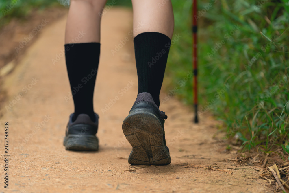 Fototapeta premium Close-up of woman leg walking on dirt trail wearing black socks and athletic shoes with hiking stick support in nature