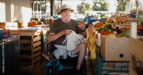 Senior man in an electric wheelchair shops for fresh vegetables at a city market, smiling and talking with a female vendor while placing tomatoes into a reusable linen bag.