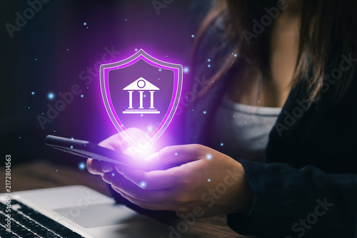 Businesswoman using smartphone with glowing bank security shield icon, illustrating secure mobile banking, financial protection, digital payment, fintech and online account safety.