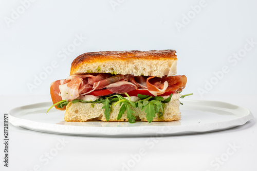 Gourmet Italian prosciutto sandwich filled with fresh mozzarella, ripe tomatoes, and arugula on crusty focaccia bread, served on a white plate in studio light.