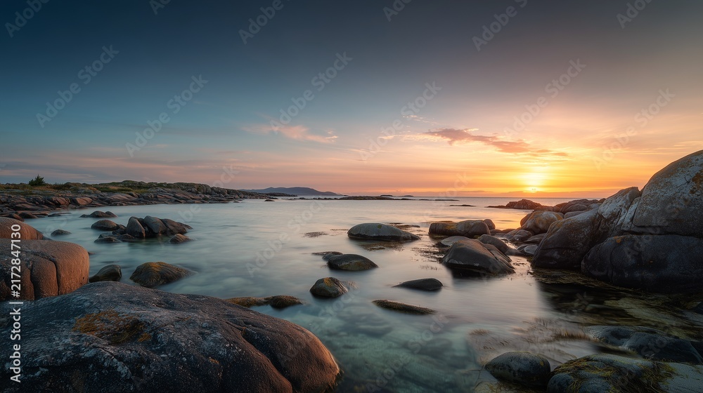 Obraz premium Majestic golden sunset illuminates calm coastal waters featuring smooth grey boulders scattered along the shoreline under a dramatic soft twilight sky during a peaceful summer evening