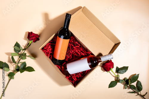 Close up of a gift box with two wine bottles and a red rose resting on red paper shred. Romantic date night or anniversary gift delivery.
