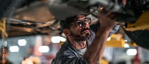 Automotive mechanic inspecting vehicle undercarriage in repair garage with focused expression and protective eyewear during maintenance service