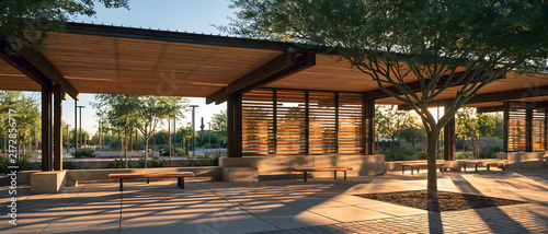 Modern outdoor pavilion with wooden slat walls, shaded seating, mature tree, and warm sunlight creating patterned shadows across landscaped public plaza