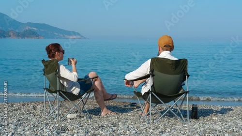 Couple relaxing on beach chairs, enjoying tranquil ocean view. Tourists enjoy a peaceful moment on a pebble beach, seated in camping chairs, sipping from mugs and gazing at the serene blue waters