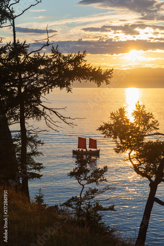 Baikal Lake at summer sunset. Silhouettes of coastal larches of Olkhon Island against setting sun. Group of tourists travel on sailboat and admire beautiful sunset. Scenic landscape. Travels, outdoors