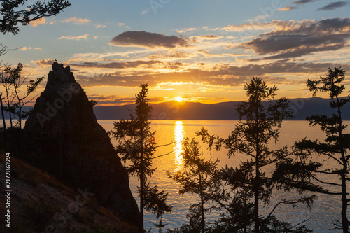 Lake Baikal at sunset in summer. Silhouettes of coastal larch trees of Olkhon Island against backdrop of setting sun on mountains. Picturesque landscape. Nature park. Travel and outdoor recreation