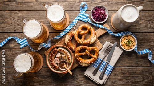 Overhead shot of a traditional snack board featuring soft pretzels, sliced sausages, mustard, and frothy beer on a wooden table.