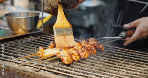 Chef brushing sauce on meat skewers  grilling on outdoor street food stall  Asian night market culinary experience  professional cooking process.