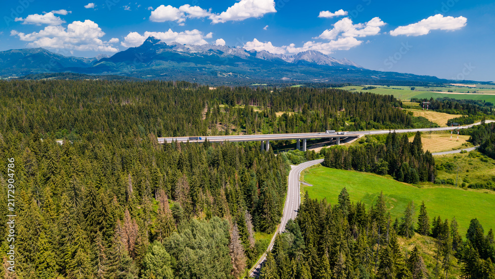 Fototapeta premium Winding road and mountains in the background during daytime. A winding road weaves through green fields and forests, mountains looming in the distance.
