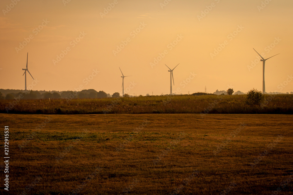 Obraz premium Modern wind turbines silhouette in agricultural field against warm hazy orange sunset sky