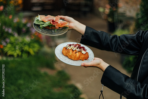 Person holds two plates with an open prosciutto and leafy green sandwich plus a cream-topped croissant, photographed outdoors with soft garden bokeh and copy space