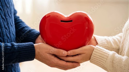 Close-up of a large red heart-shaped piggy bank held by a man and a woman from both sides. Building long-term relationship, financial unity and lifelong experience in long-term marriage concept