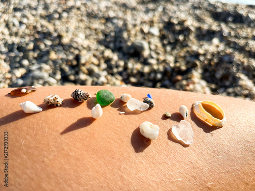 Close-up of small sea shells and colorful polished sea glass pieces arranged in a row on a suntan skin over a pebble beach background